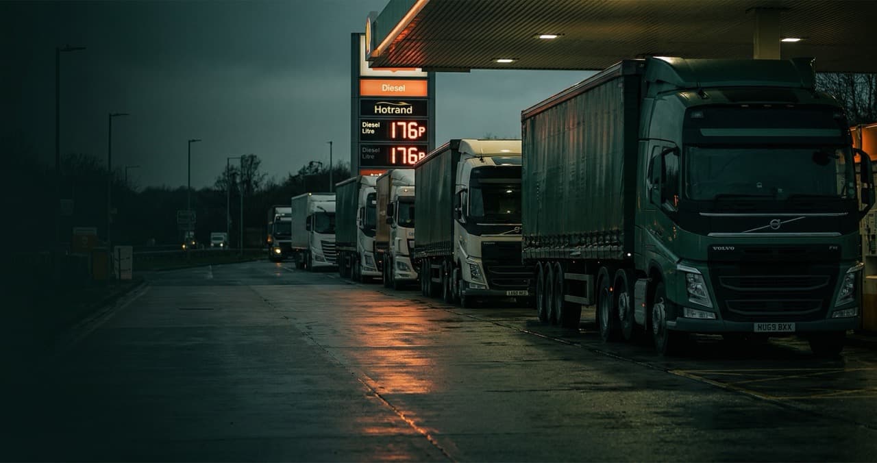 Row of HGV lorries queuing at a UK motorway diesel forecourt at dusk, fuel price board visible in background